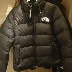 Svart pufferjacka från The North Face - Säljer en snygg och varm svart pufferjacka från The North Face. Jackan har en dragkedja framtill och är perfekt för kalla vinterdagar. Den är i bra skick och har en klassisk design med loggan på bröstet och ryggen. Perfekt för både vardag och utomhusaktiviteter!