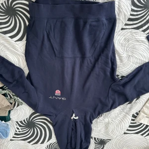 Mörkblå hoodie från GANT - Säljer en snygg och bekväm mörkblå hoodie från GANT. Den har en klassisk känguruficka och en justerbar dragsko i huvan. Perfekt för en avslappnad stil eller för att hålla sig varm under kyliga dagar. GANT-loggan är broderad på framsidan. Passar perfekt till jeans eller joggers!