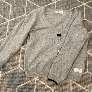 Grå kofta från Odd Molly - Säljer en superfin grå kofta från Odd Molly. Den är i en mjuk och skön kvalitet med en söt liten svart rosett vid knäppningen. Koftan har en v-ringad design och knappar framtill. Perfekt för både vardag och lite finare tillfällen!