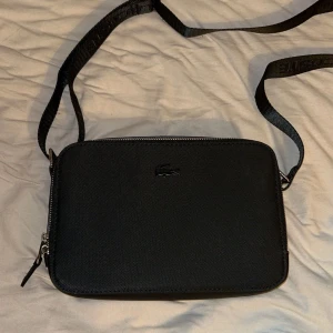Lacoste Väska - Stilren svart Lacoste väska i utmärkt skick. Perfekt för att förvara dina tillhörigheter