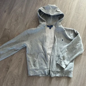 Polo Ralph Lauren hoodie - Säljer en snygg och bekväm grå hoodie från Polo Ralph Lauren. Den har en dragkedja framtill och två praktiska fickor. Perfekt för en avslappnad stil och passar till de flesta tillfällen. Den är i bra skick och redo för en ny ägare!