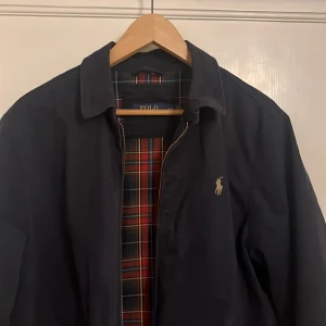 Mörkblå jacka från Polo Ralph Lauren - Snygg mörkblå jacka från Polo Ralph Lauren. Den har en klassisk design med dragkedja och ett stilrent rutigt foder i rött och grönt. Jackan är perfekt för höst och vår, och har en broderad logga på bröstet. Den är i nyskick och passar perfek