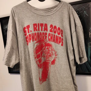 Grå t-shirt med röd text - Säljer en grå t-shirt med röd text 'ST. RITA 2008 SOPHOMORE CHAMPS' och en cool grafik av en hjälm. Den är kortärmad och perfekt för en avslappnad stil. Passar bra till jeans eller shorts!