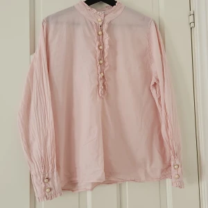 Rosa blus med pärlknappar - Säljer en superfin rosa blus med långa ärmar och vackra pärlknappar. Blusen har en söt volangdetalj längs knäppningen och är perfekt för både vardag och fest. Den är i bra skick och redo för en ny ägare!