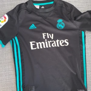 Svart Real Madrid fotbollströja från Adidas - Säljer en svart Real Madrid fotbollströja från Adidas med korta ärmar och turkosa detaljer. Tröjan har klubbens emblem och Fly Emirates-logga på framsidan. Perfekt för fans av laget! 🖤