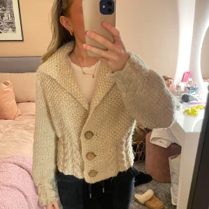Beige handstickad kofta med knappar - Mysig beige stickad kofta med stora träknappar framtill. Koftan har en grovstickad struktur och en bred krage som ger en avslappnad look. Den är i väldigt bra kvalitet och är i ull. Den är varm. Det är ingen storlek men skulle säga ca S.