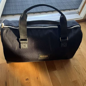 Svart väska från Boss - Snygg svart väska från Boss med praktiska handtag och dragkedja. Perfekt för att bära med sig det nödvändigaste. Stilren design med märkets logga på framsidan.