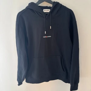 Svart hoodie från Saint Laurent size L - Säljer en stilren svart hoodie från Saint Laurent med logotyp på bröstet. Tröjan har en klassisk design med huva och dragsko samt en praktisk magficka. Perfekt för en avslappnad look. Passar längder mellan 180-190 cm inga defekter väldigt bra skick 