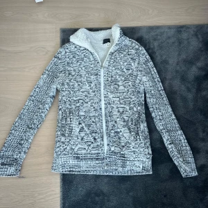 Stickad cardigan - Säljer en grå stickad cardigan från Jeel med dragkedja och mysigt foder. Tröjan har ett snyggt mönster och långa ärmar, perfekt för kyliga dagar. Den har en hög krage som ger extra värme och stil. Passar bra till både jeans och casual outfits. Riktigt fet och riktigt skön! 