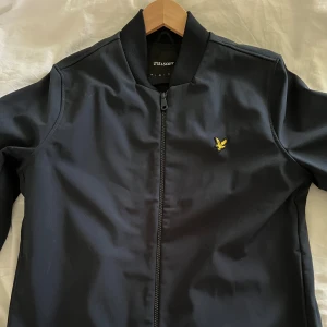 Svart bomberjacka från Lyle & Scott - Snygg mörkblå bomberjacka från Lyle & Scott med dragkedja och ett litet broderat emblem på bröstet. Jackan är tillverkad i polyester (softcell jacka) och har långa ärmar. Perfekt för en stilren look.