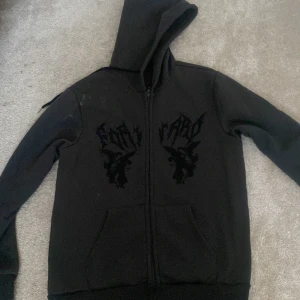 Svart hoodie med tryck - Säljer en svart hoodie med dragkedja och tryck på bröstet. Den har en klassisk huva och långa ärmar. Perfekt för en avslappnad stil.