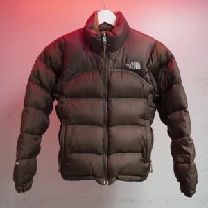 Brun dunjacka från The North Face - Säljer en brun dunjacka från The North Face med dragkedja och hög krage. Jackan har långa ärmar och justerbara ärmslut. Perfekt för kalla dagar och håller dig varm och bekväm. 🧥