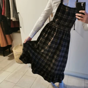 Rutig klänning med volangdetalj - Charmig rutig klänning i mörka toner med volangdetalj vid fållen. Egentligen är det lite mer av ett förkläde, men man kan absolut ha den som en klänning. Klänningen har en klassisk passform med knytband i midjan och band som man knyter bakom halsen.. Längd: 107 cm. 