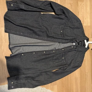 Mörkgrå skjorta från Indicode Jeans - Snygg mörkgrå och trendig skjorta från Indicode Jeans med knappar framtill och två bröstfickor. Perfekt för en stilren look. Skjortan har långa ärmar och en klassisk krage. Passar även som M. Skicket är 10/10 och finns inga defekter