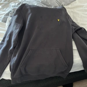 Svart hoodie från Lyle & Scott - Säljer en stilren svart hoodie från Lyle & Scott Junior. Den har en klassisk design med en känguruficka framtill och en liten gul logga på bröstet. Perfekt för en avslappnad look.