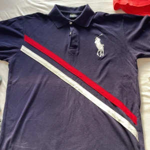 Mörkblå pikétröja från Ralph Lauren - Säljer en snygg mörkblå pikétröja från Ralph Lauren med klassisk krage och knappar. Tröjan har ett stort vitt broderat logotypmotiv på bröstet och diagonala ränder i rött och vitt. Tillverkad i 100% bomull för en bekväm passform.