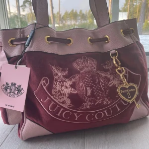 Lila/Rosa och beige väska från Juicy Couture - Snygg och rymlig brun och beige väska från Juicy Couture med guldiga detaljer. Väskan har ett stort huvudfack och flera innerfickor, perfekt för att organisera dina saker. Den har också ett dekorativt hjärta med ett 'J' på baksidan. Perfekt för dig som vill ha både stil och funktion. Endast andvänd ett fåtal gånger så den är så gott som ny men kan ändå finnas nån pytteliten skada jag inte märkt !första bilden är ej min, lappen är bortklippt men ligger inuti väskan!