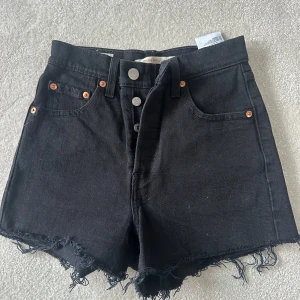 Svarta jeansshorts från Levi's - Snygga svarta jeansshorts från Levi's med hög midja och fransiga kanter. De har en knappgylf med fyra knappar och klassiska femficksdetaljer. Används fåtal gånger