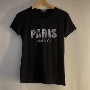 Svart t-shirt från Attitude Paris - Snygg svart t-shirt från Attitude Paris med texten 'PARIS FRANCE' i glittriga bokstäver. Perfekt för en stilren look med en touch av glamour. Kortärmad och bekväm passform. Den är storlek L men den är absolut inte så stor tycker jag.