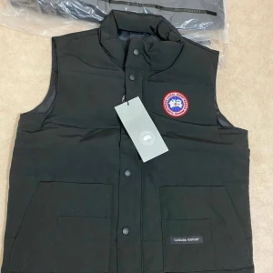 Svart dunväst från Canada Goose - Snygg svart dunväst från Canada Goose med ett ikoniskt märke på bröstet. Västen har en hög krage och knäppning framtill. Perfekt för kyliga dagar när du vill hålla värmen utan att kompromissa med stilen. Storlek M men passar S