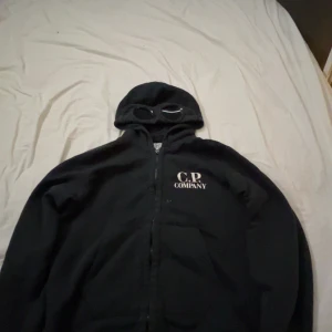Svart huvtröja med glasögon från C.P. Company - Säljer en cool svart huvtröja från C.P. Company med inbyggda glasögon i huvan. Tröjan har dragkedja framtill och är perfekt för en unik stil. Märkeslogga syns tydligt på bröstet. Passar perfekt för dig som vill sticka ut!