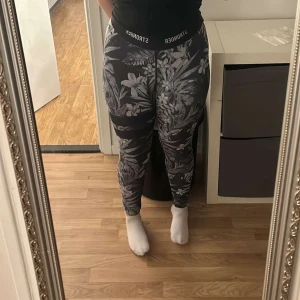 Svarta leggings med blommigt mönster från Stronger - Snygga svarta leggings från Stronger med ett stilrent blommigt mönster i grått och vitt. Perfekta för träning eller en avslappnad dag. De har en hög midja för extra komfort och stöd.