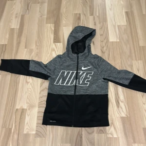 Grå och svart hoodie från Nike - Snygg grå och svart hoodie från Nike med stor logga på bröstet. Tillverkad i Dri-FIT-material för optimal komfort. Perfekt för träning eller en avslappnad stil. Har dragkedja och huva för extra skydd.