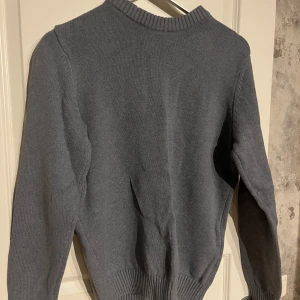 Grå stickad tröja - Säljer en stilren grå stickad tröja med ribbad krage och muddar. Perfekt för kyligare dagar och enkel att matcha med olika outfits. Tröjan har långa ärmar och en klassisk passform.