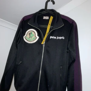 Svart zip dress från Palm Angels x Moncler - Säljer en svart zip dress från Palm Angels i samarbete med Moncler. Jackan har en dragkedja framtill och ett stort Moncler-märke på bröstet. Den har långa ärmar med en lila rand längs sidan. Perfekt för dig som gillar stilrena och sportiga plagg.