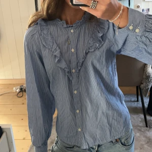 Blå randig blus med volangdetaljer - Säljer en söt blå randig blus med volangdetaljer längs kragen och bröstet. Blusen fungerar till både finare tillställningar och till vardags! Jättefin till sommaren!🌟Använd endast 1 gång, köpt för 400kr