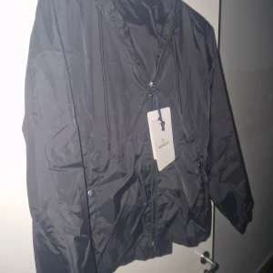 Svart vindjacka från Moncler Windbreaker - Snygg svart vindjacka från Moncler med dragkedja och huva. Jackan har Moncler-logga på ärmen och justerbara snören vid huvan. Perfekt för blåsiga dagar.