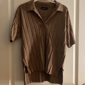 Brun ribbad tröja från KappAhl - Säljer en brun ribbad tröja från KappAhl med korta ärmar och en klassisk krage. Perfekt för en stilren look. Tröjan har en avslappnad passform och är tillverkad i ett mjukt material.
