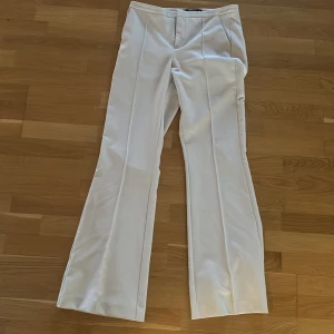 Begia kostymbyxor - Eleganta begia kostymbyxor med låg midja och bootcut-stil. Perfekta för en stilren look. Byxorna har en klassisk design med pressveck och stängs med dragkedja och knapp.