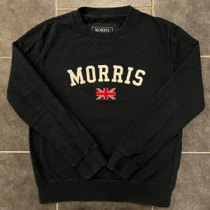 Svart sweatshirt från Morris - Säljer en stilren svart sweatshirt från Morris med broderad text och en brittisk flagga på bröstet. Tröjan har långa ärmar och ribbade muddar vid ärmslut och nederkant. Perfekt för en avslappnad stil.