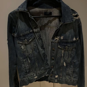 Jeansjacka - Säljer en blå jeansjacka med slitningar och knappar framtill. Jackan har två bröstfickor med lock och knappar, samt långa ärmar. Perfekt för vår och höst men även sommar🥰 