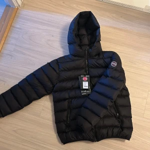 Svart dunjacka från Colmar - Snygg svart dunjacka från Colmar med huva och dragkedja. Jackan har en quiltad design och en rund logotyp på ärmen. Perfekt för kyliga dagar.