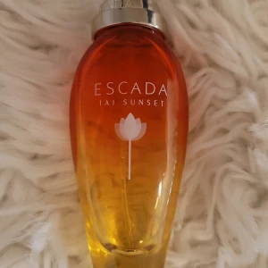Escada Taj Sunset Parfym - Säljer en Escada Taj Sunset parfym i en elegant flaska med en gradient från orange till gul. Flaskan har en silverfärgad kork och en stilren design med en vit blomma på framsidan. Perfekt för den som älskar fruktiga och exotiska dofter. 30 ml, se andra bild för att se kapasitet