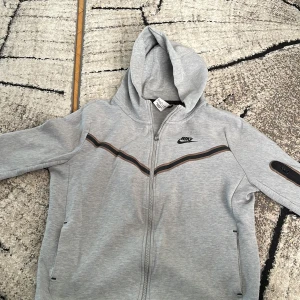  Nike take - Säljer en grå huvtröja från Nike med dragkedja och huva. Tröjan har en svart och brun randdetalj över bröstet och en ficka på ärmen. Perfekt för en sportig look eller avslappnade dagar.