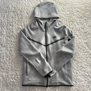 Grått mjukisset med hoodie och byxor från Nike - Säljer ett stilrent set med en grå hoodie och gråa byxor från Nike med dragkedja och huva. Perfekt för en avslappnad look. Hoodien har långa ärmar med en ficka på ena ärmen och en diskret Nike-logga på bröstet. Byxorna har en logga på högra sidan och även en lång dragkedja på sidan. Passar bra till både träning och vardag.