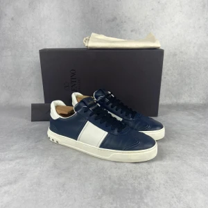 Valentino sneakers i blått och vitt - Snygga Valentino sneakers i en stilren kombination av blått och vitt. Skorna har en klassisk design med snörning och en vit sula. Perfekta för en trendig och avslappnad look.