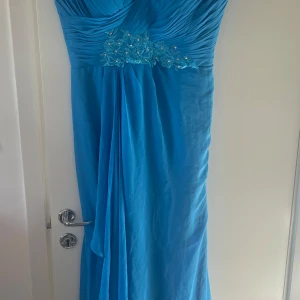 Blå axelbandslös klänning 2st 200kr st - Säljer en vacker blå axelbandslös klänning med draperad överdel och dekorativa pärlor vid bysten. Perfekt för festliga tillfällen. Klänningen har en elegant fall och en lång design som ger en sofistikerad look.