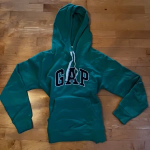 Grön hoodie från GAP - Säljer en snygg grön hoodie från GAP med stor logga på bröstet. Den har en justerbar huva! Storleken är M men skulle säga att den passar S-XS! Hör av er vid frågor😊