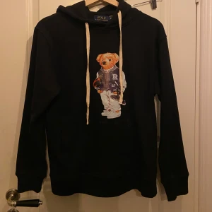 Svart polo bear från polo Ralph Lauren  - Säljer en svart hoodie från Polo Ralph Lauren med  polo björnen på framsidan. Hoodien har långa ärmar och en justerbar huva med vita snören. Perfekt för en avslappnad stil.