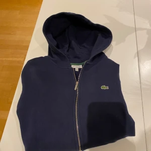 Mörkblå hoodie från Lacoste - Säljer en snygg mörkblå hoodie från Lacoste med dragkedja och huva. Den har en klassisk krokodil-logga på bröstet och praktiska fickor framtill. Perfekt för en avslappnad stil.
