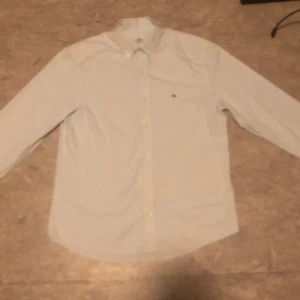 Vit skjorta från Lacoste - Säljer en stilren ljusblå skjorta från Lacoste med broderad logga på bröstet. Skjortan har en slim passform och långa ärmar med knappar vid ärmsluten. Perfekt för en snygg och klassisk look.