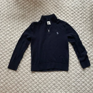 Mörkblå halfzip från Abercrombie & Fitch - Snygg mörkblå halfzip tröja. Perfekt för kyligare dagar. Den har långa ärmar och en liten broderad logga på bröstet. Ribbstickade muddar vid ärmslut och nederkant ger en stilren look. 