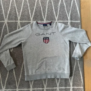 Grå sweatshirt från GANT - Säljer en stilren grå sweatshirt från GANT med ett broderat emblem på bröstet. Tröjan har långa ärmar och en rund halsringning med ribbade muddar. Perfekt för en avslappnad stil. Om ni undrar något är det bara att kontakta mig
