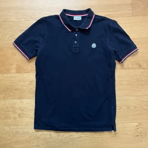 Mörkblå pikétröja från Moncler - Snygg mörkblå pikétröja från Moncler med klassisk krage och knappar. Tröjan har röda och vita detaljer på kragen och ärmsluten samt Monclers logga broderad på bröstet. Perfekt för en stilren look. Du vet varför så den är så billig 🐼. Den är i bra skick. Jag rekommenderar den starkt till någon som vill ha lite extra fina kläder till sommaren! Mitt pris är fast på 199kr förutom om man är en följare då får man den för 179kr! Ställ gärna frågor!