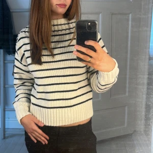 Vit och svart randig stickad tröja - Säljer en snygg vit stickad tröja med svarta ränder. Tröjan har ”boat neck” ribbad krage och långa ärmar, perfekt för kyligare dagar. Den har en avslappnad passform och är supermjuk och bekväm. Perfekt för en stilren look!