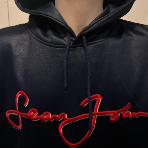 Mörkblå hoodie från Sean John - Säljer en snygg mörkblå hoodie från Sean John med röd broderad logga på framsidan. Tröjan har en klassisk känguruficka och ribbade muddar med röd och vit detalj vid ärmslut och nederkant. Perfekt för en avslappnad stil.